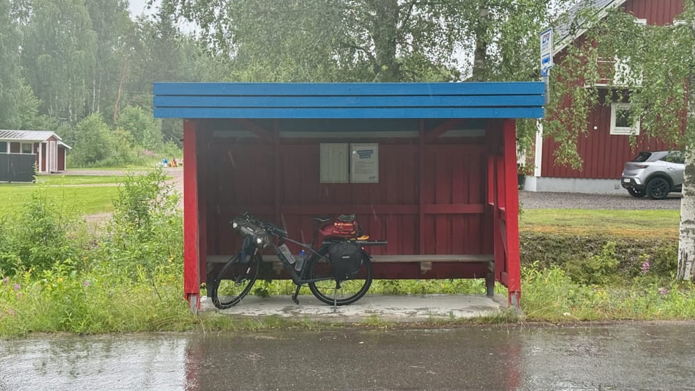 A bicycle is locked onto a red bench on a street and it is raining.