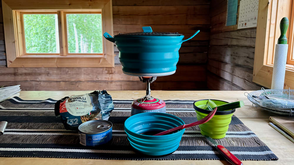 A camping stove sits on a table next to some containers with food in them and a blue container with a handle.
