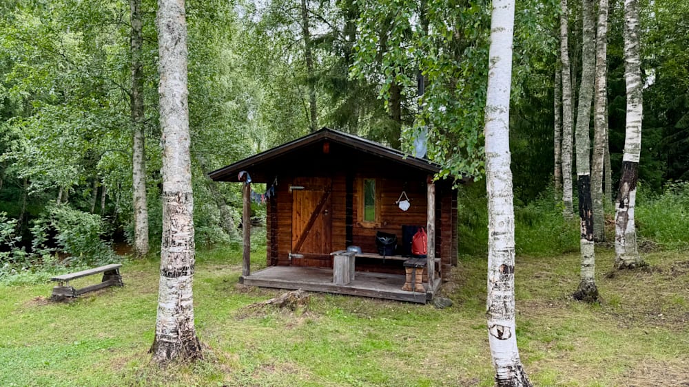 A small cabin in the woods with a bench outside and a backpack on the porch.