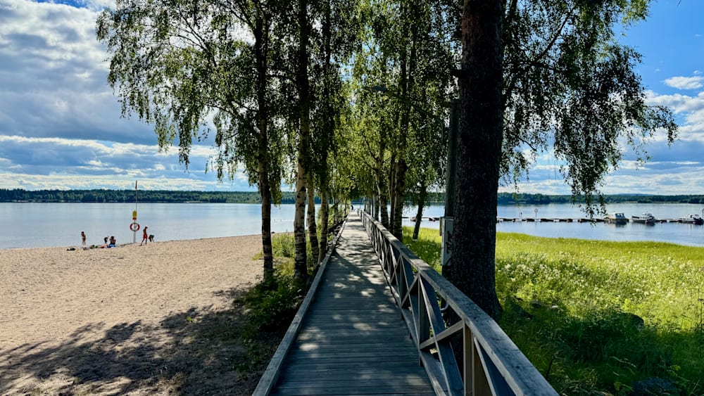 A long boardwalk that goes along a lake with trees and a bridge in the foreground.