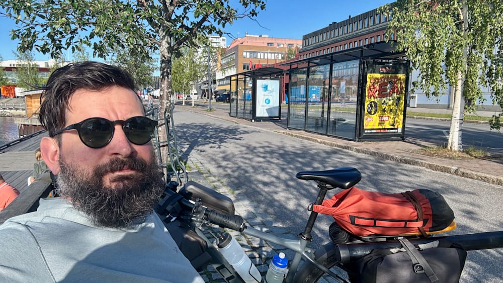 A man wearing sunglasses sits on a bicycle next to a bench.