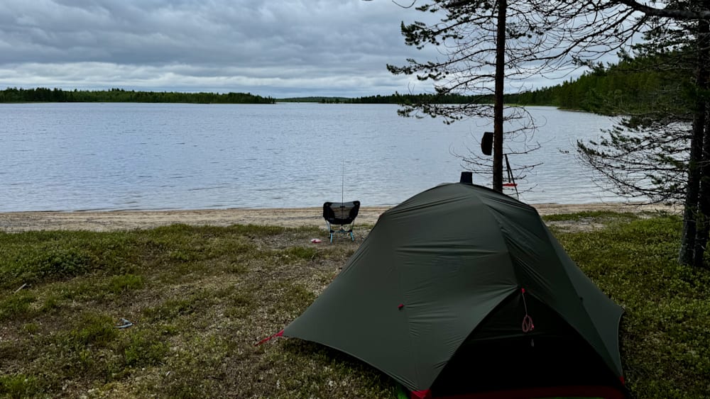 A tent on a beach next to a lake with trees in the background.