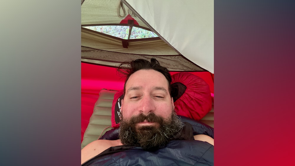 A man with a beard and a red pillow laying in a tent.