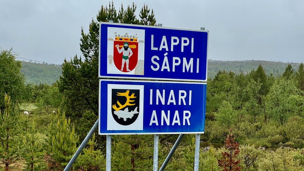 A blue sign with two signs on it in front of trees.