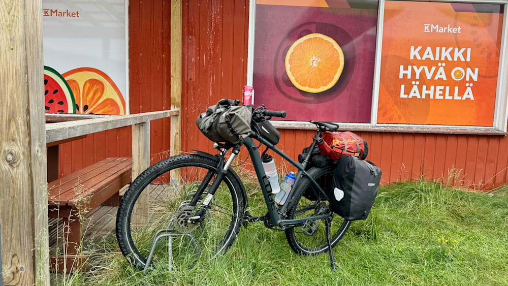 A bike with a bottle of water and a bag on the back of it parked next to a red building with an orange fruit print.