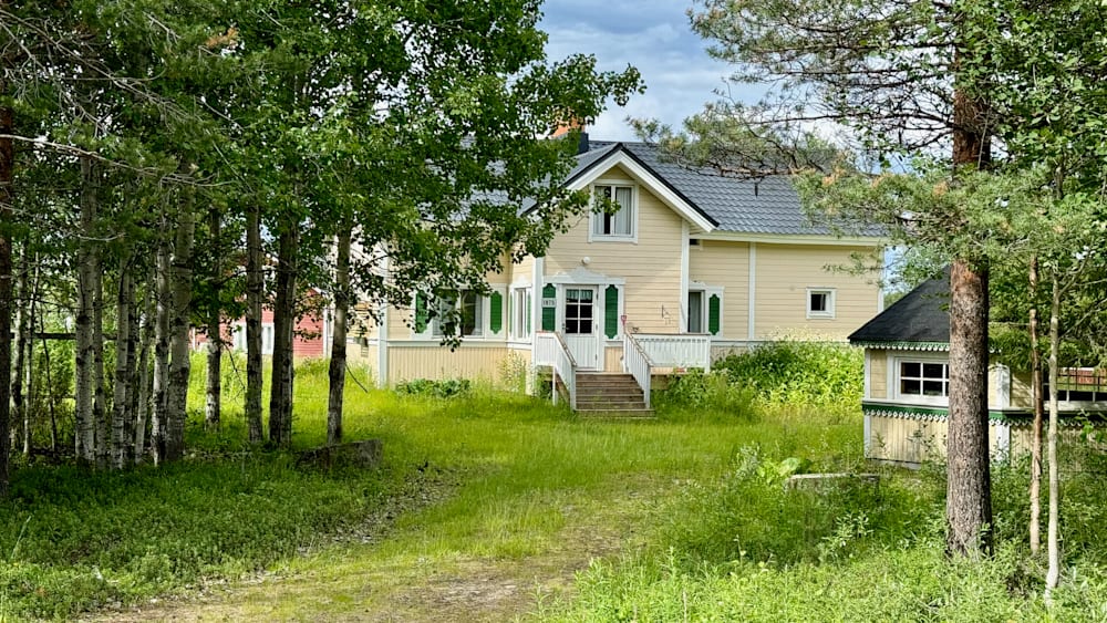 A large yellow house with green shutters is shown on a tree covered street.