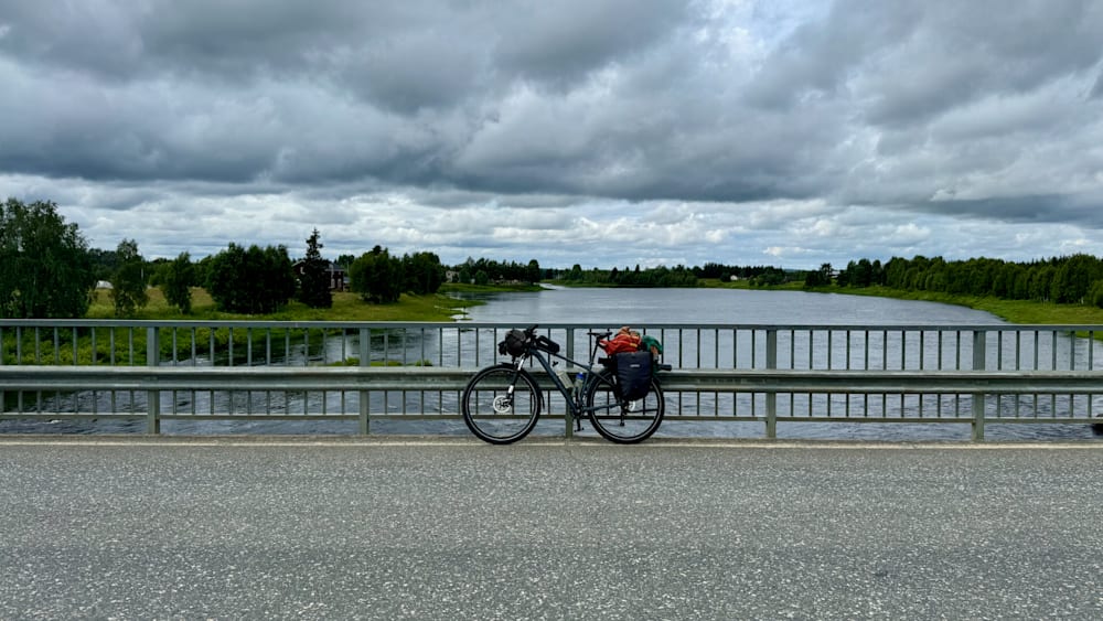 A bike is locked onto a railing and sits on a road next to a bridge.