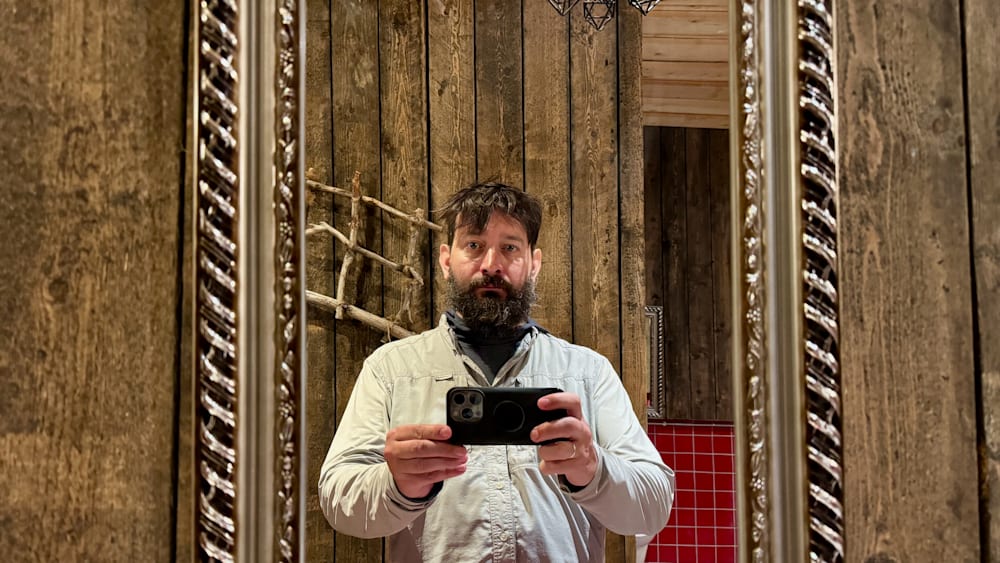 A man with a beard and a white shirt is holding up his phone in front of a mirror.