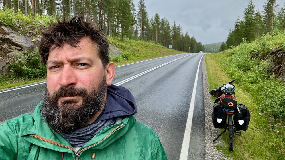 A man with a beard and green jacket stands on the side of a road.