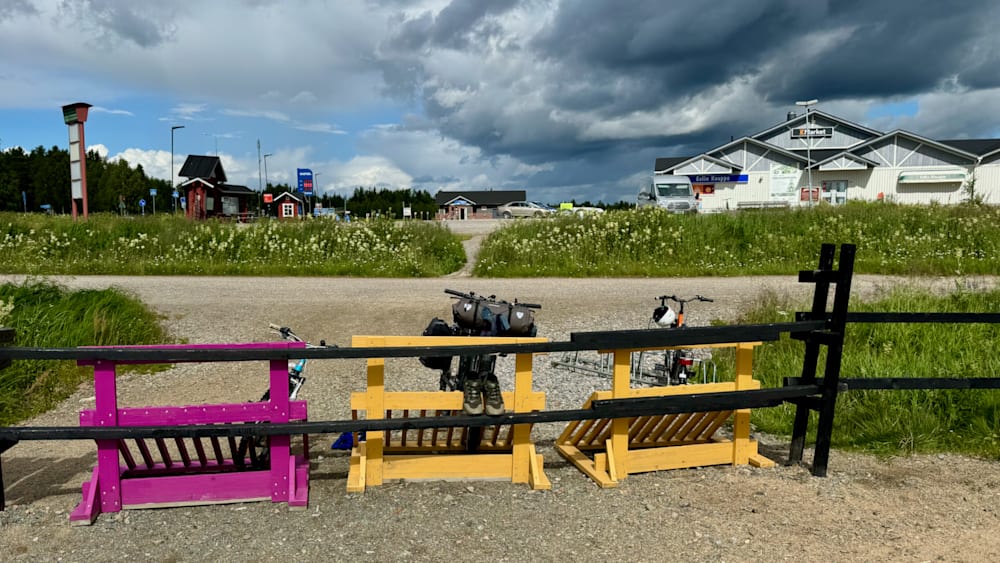 A row of bikes are leaning against a fence on a gravel lot.