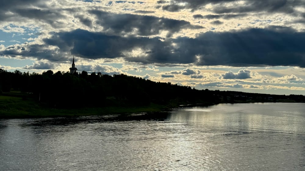 A river bank is shown with a church in the background under a cloudy sky.