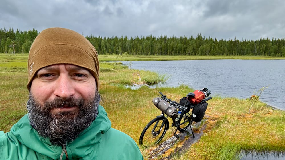 A man wearing a green jacket takes a picture of himself with a bicycle that is parked next to him.