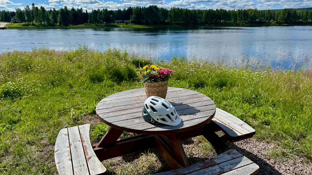 A picnic table with flowers and a helmet on it by the water.