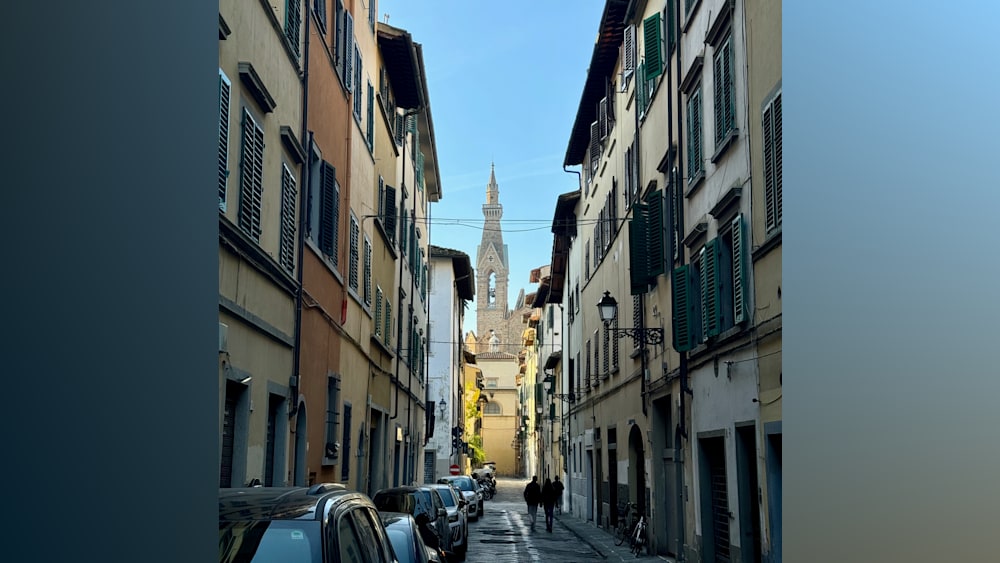 A narrow, brick city street with several cars parked on both sides and a tall, spire-like building in the background.