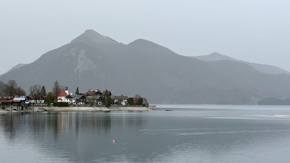 A boat is on the water in front of a small town with mountains in the background.