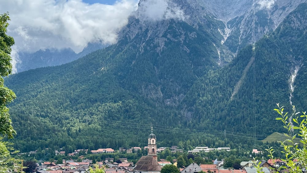 A church with a spire on a mountain with trees and a town in the valley below.
