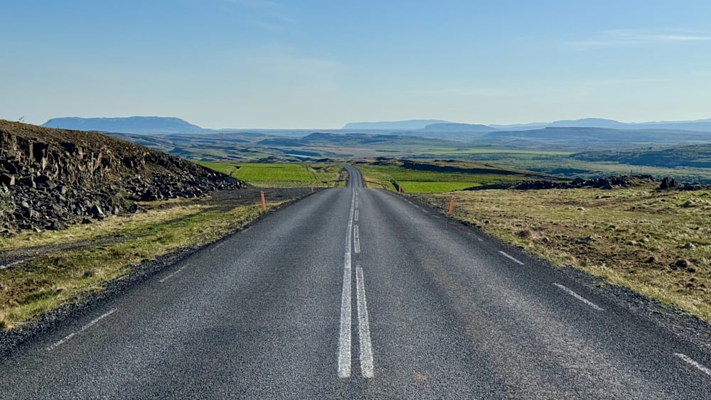 An empty road with white stripes is depicted with a large hill in the background.