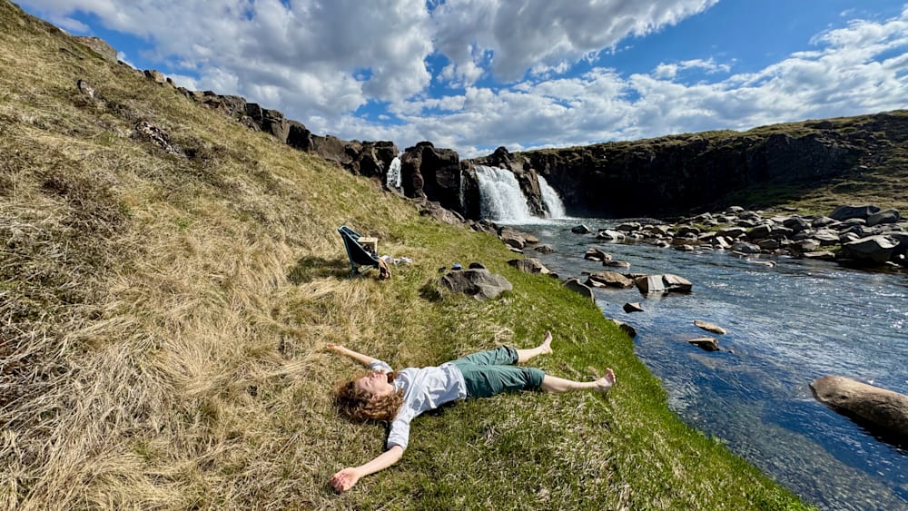 A woman is laying on the grass next to a body of water, with her back to the camera.