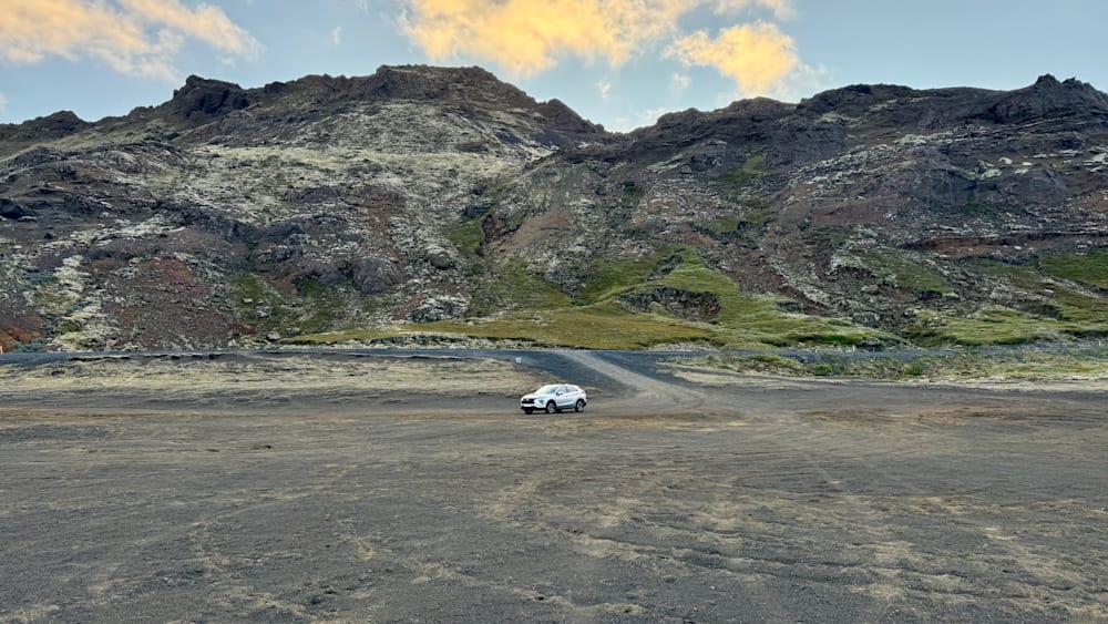 A white car is parked on a dirt road in front of a mountain range.