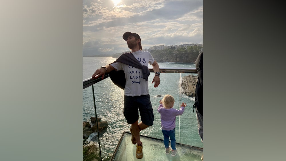 A man wearing a t-shirt that says "cycle arts labor" stands on a balcony overlooking the ocean.