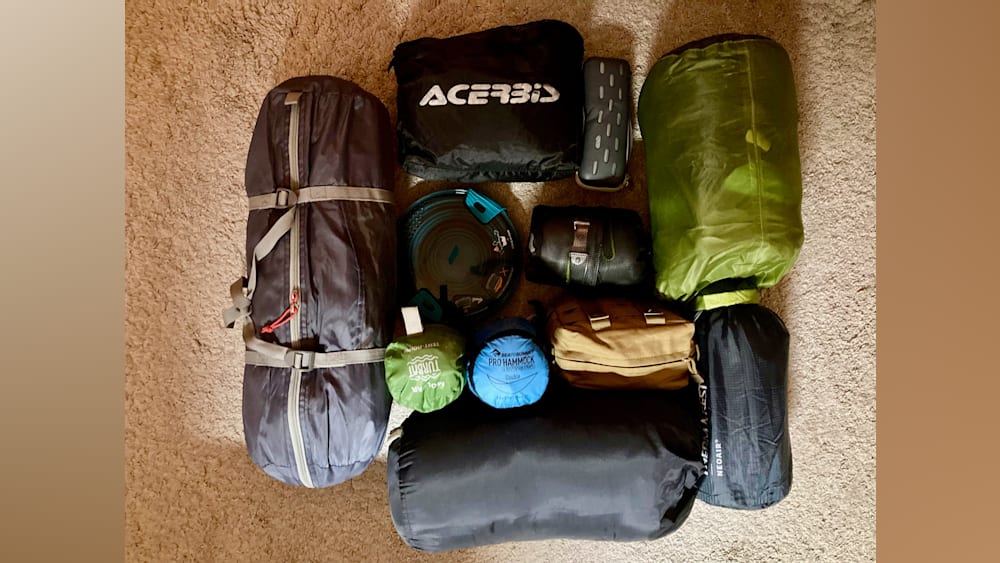 Several pieces of camping gear are spread out on a carpeted floor, including an Acecobis bag.