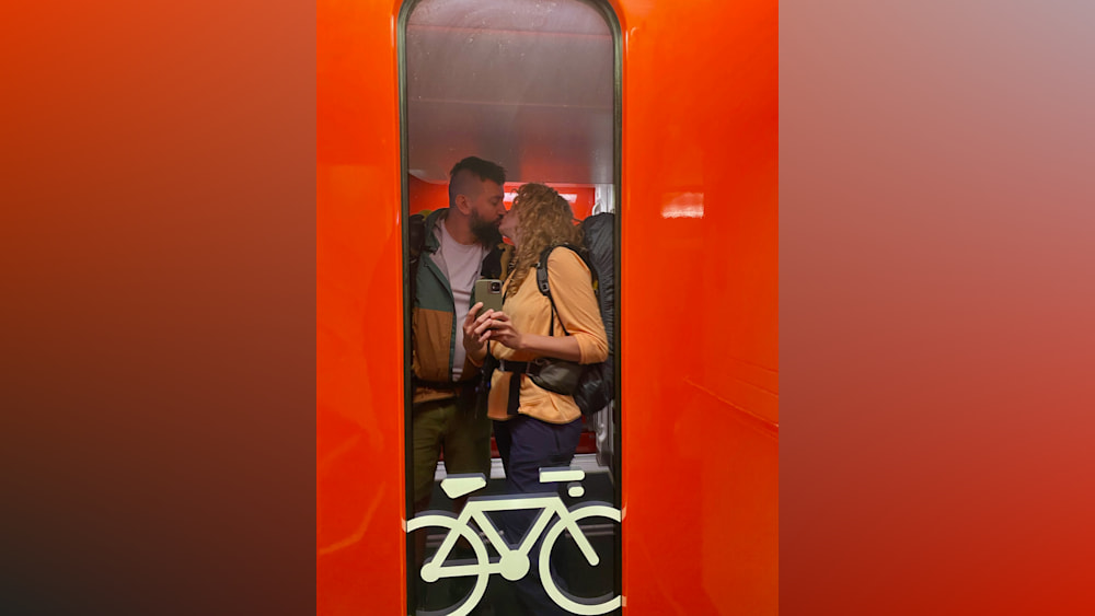 A man and woman kissing in a room with a bike and back pack in the background.