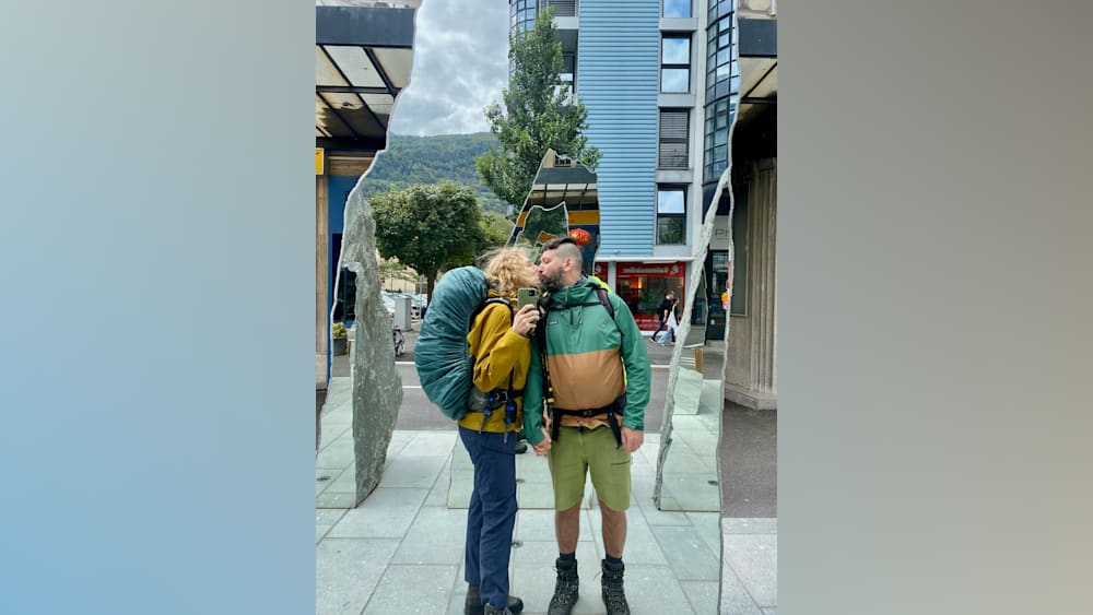 A man and woman in front of a mirror with a backpack on the woman and a camera in the man's hand.