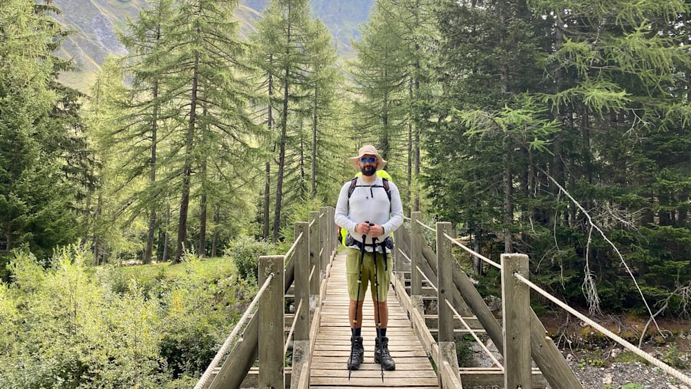 A man wearing a hat poses for a picture on a wooden bridge over a river.