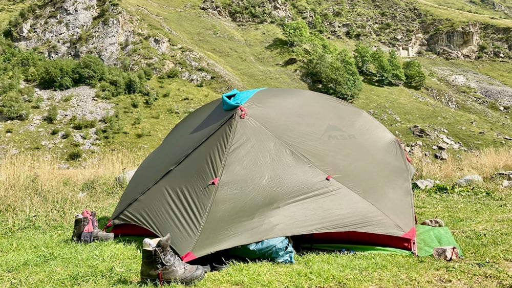 An MSR tent is set up on a grassy knoll with a pair of hiking boots beside it.