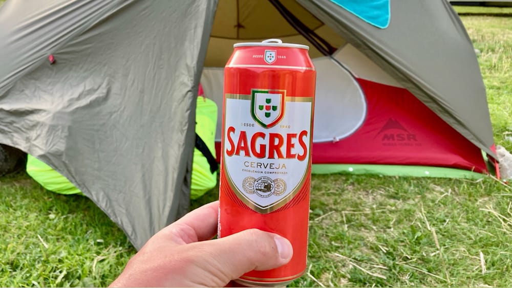 A hand holding a can of Sagres beer in front of a tent.