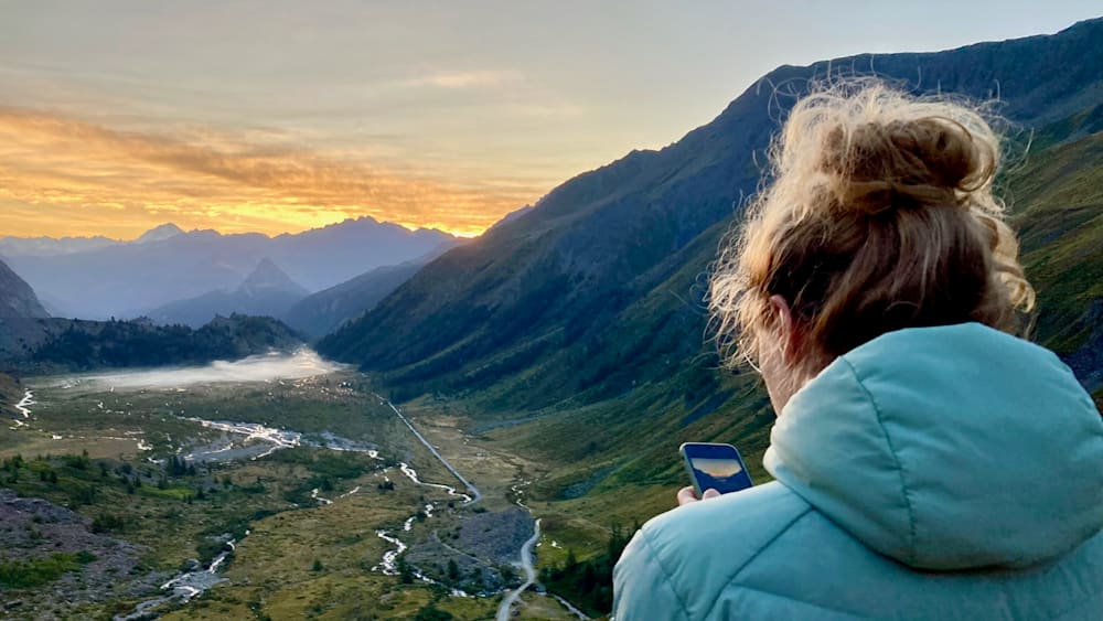 A woman in a blue jacket is looking at her phone at the edge of a mountain top.