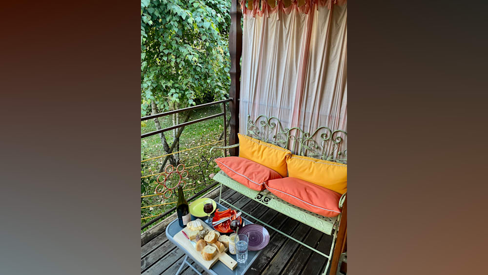 Two colorful pillows on a wrought iron bench in front of a table with wine glasses and plates of food.