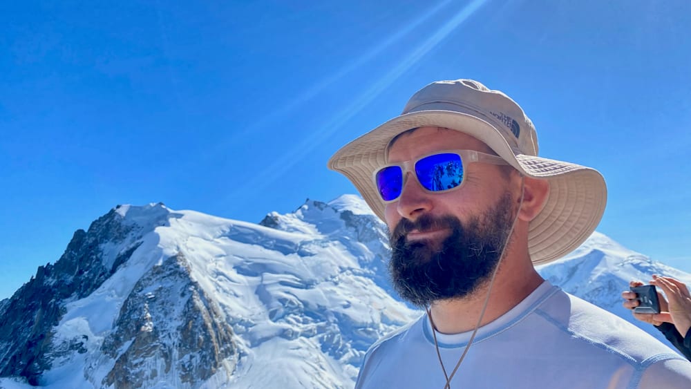 A man wearing a hat and sunglasses, with a mountain in the background.
