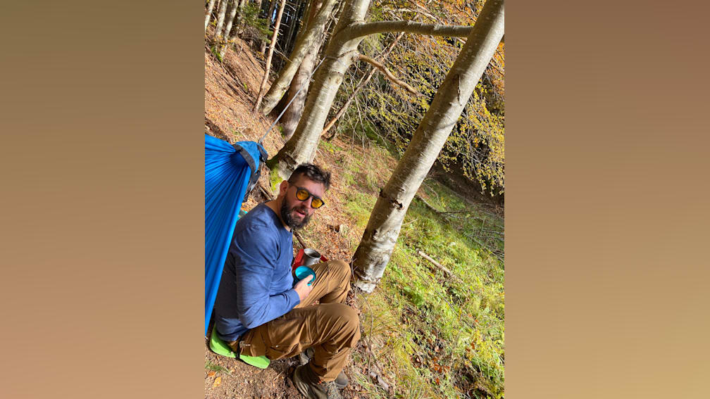 A man in a blue hammock is sitting near a tree and has a cup in his hand.