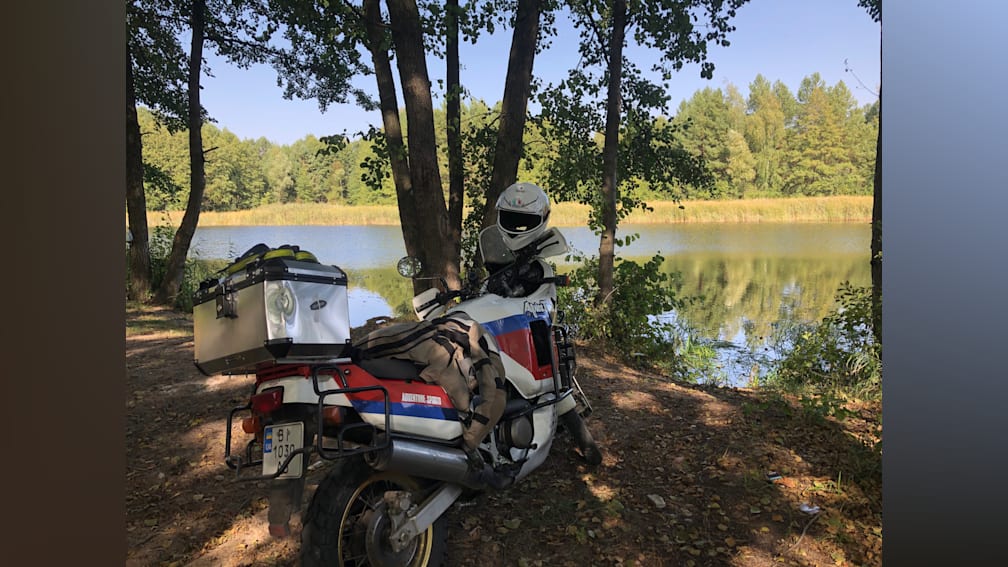 A motorcycle with a helmet on it parked next to a tree by a lake.