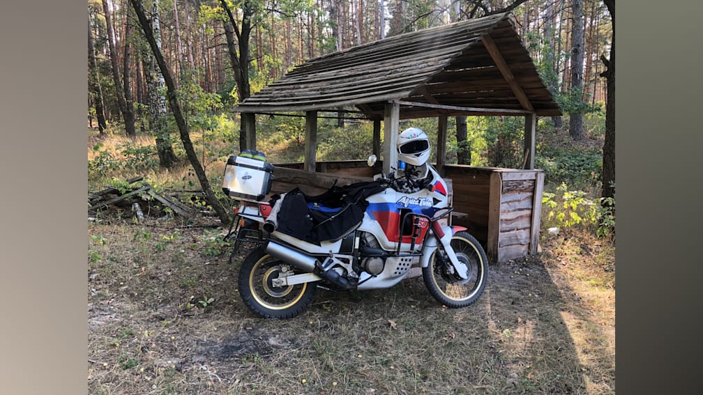 A person wearing a white helmet is sitting on a motorcycle that is parked next to a wooden structure.