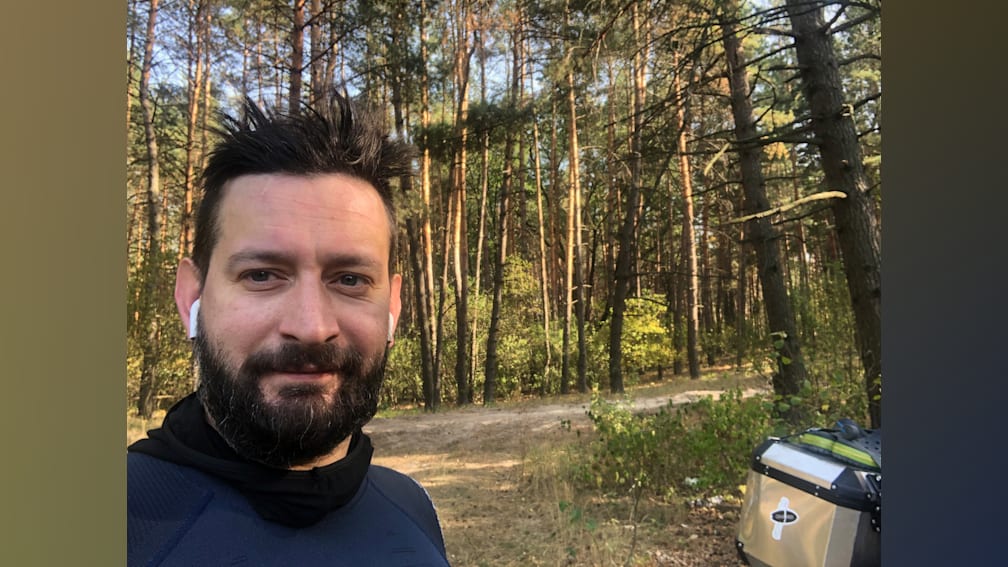 A man wearing a blue shirt is standing in front of a forest.
