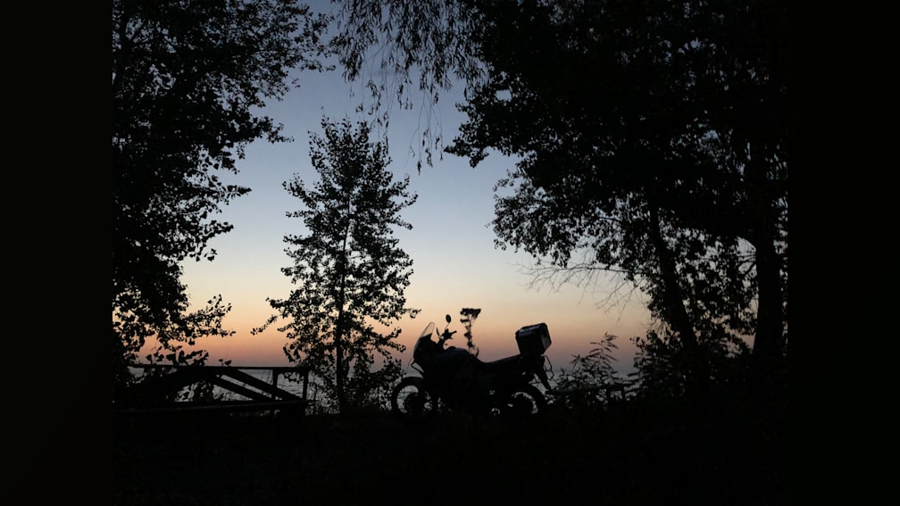 A motorcycle is parked in front of a tree line at sunset with the sun going down behind the trees.
