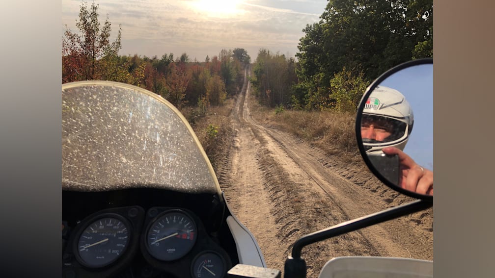 A dirt road has a gauge on a motorcycle in front of a mirror that is being used by another motorcycle rider.