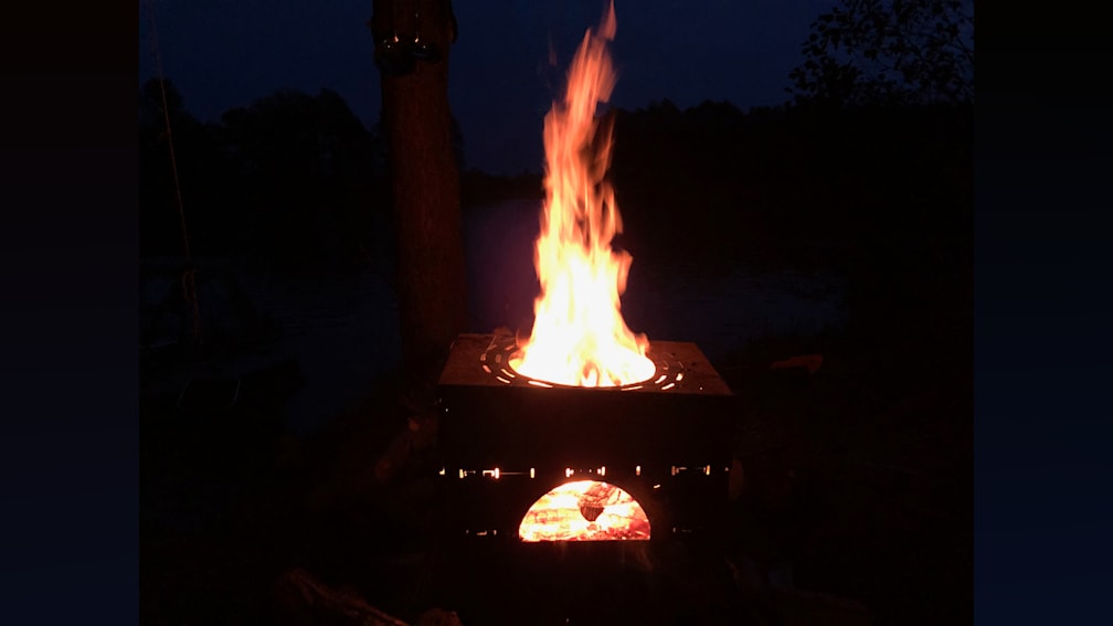 A small outdoor fire pit is lit, with a pizza cooking in it, and a tree in the background.