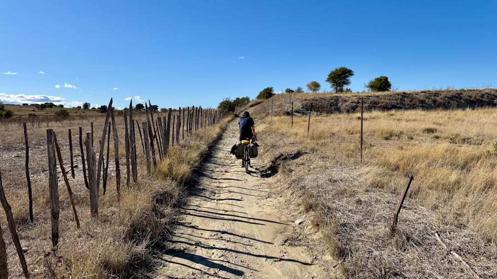 A man is riding his bike down a dirt road with his bags.