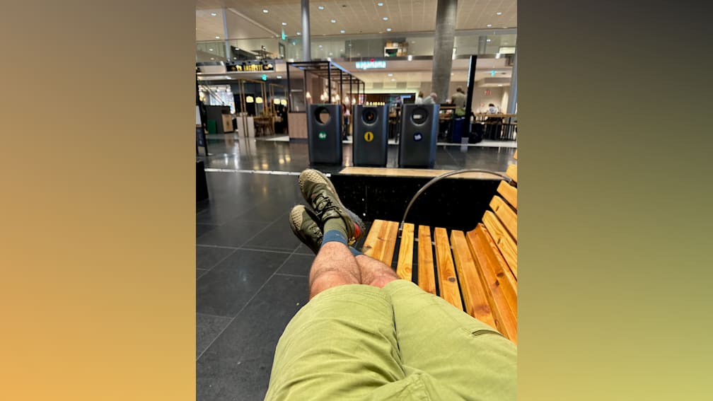 A man in a green shirt is sitting on a wooden bench with his legs up in the air.