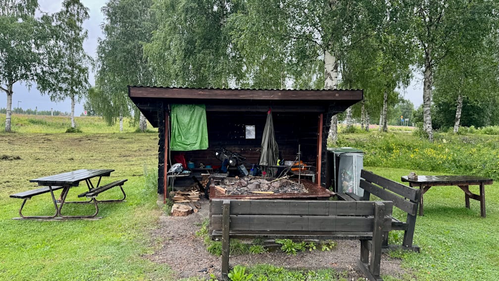 A rustic wood shelter with a green cover on a grassy area with a bench nearby.