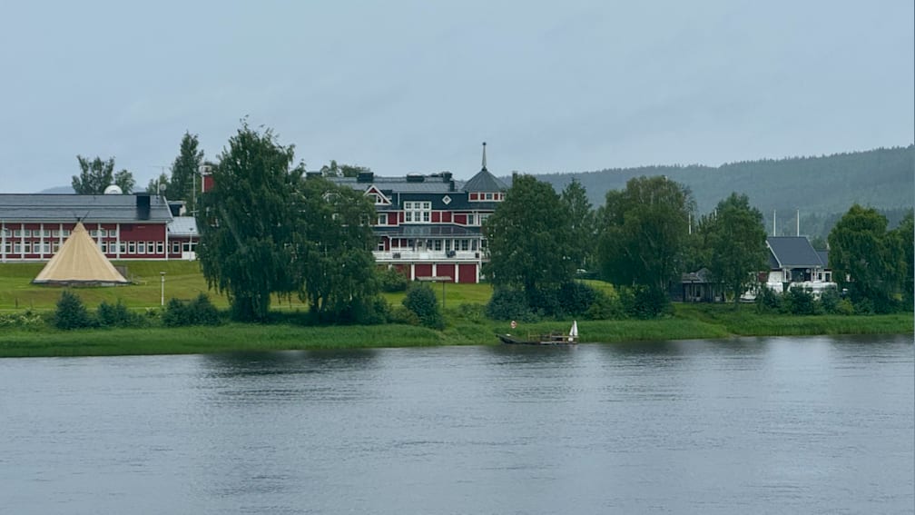 A long boat on a body of water with a red and white house on the shore.