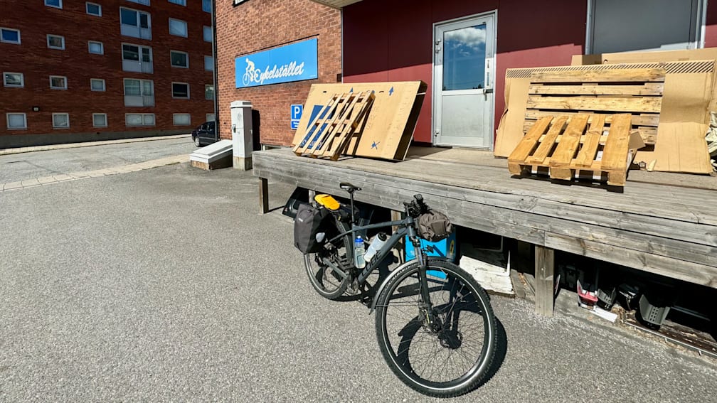 A bicycle is parked on a porch in front of a wood structure and next to a sign that says "pesistertall".
