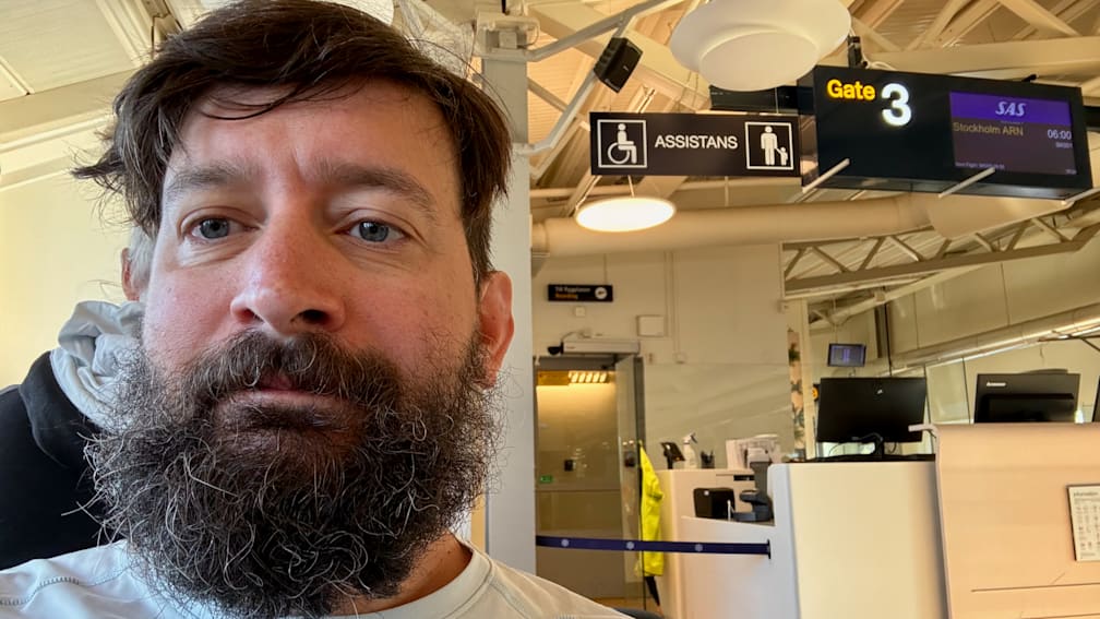 A bearded man looks at his camera as he stands in an airport with a sign that says Assistants.