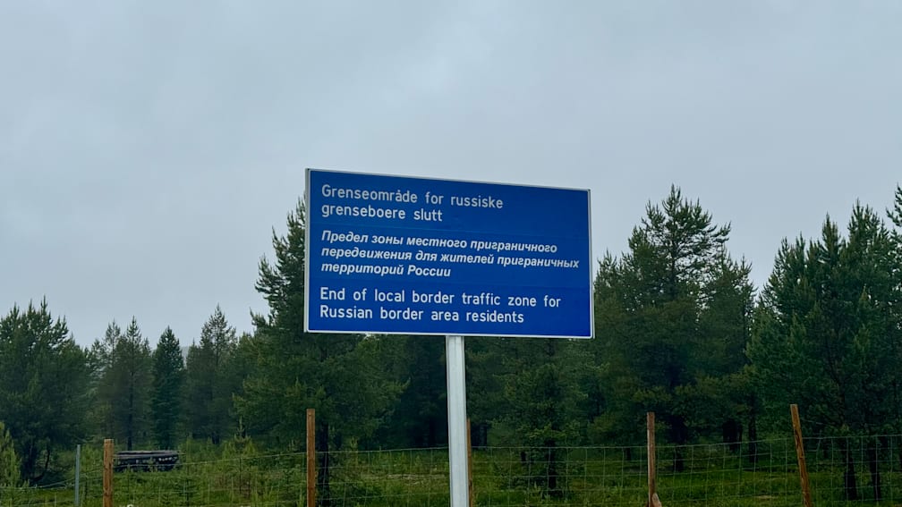 A blue sign that says "Grenesomade forRussiske Slut" is on a white pole in front of a forest.