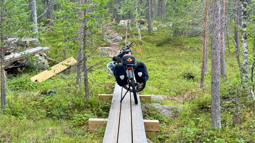 A bicycle with a black front tire and a backpack on the back is stopped on a bridge over a river.