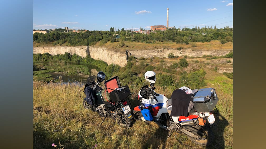 A couple on motorcycles on a grassy hill with a cliff in the background.