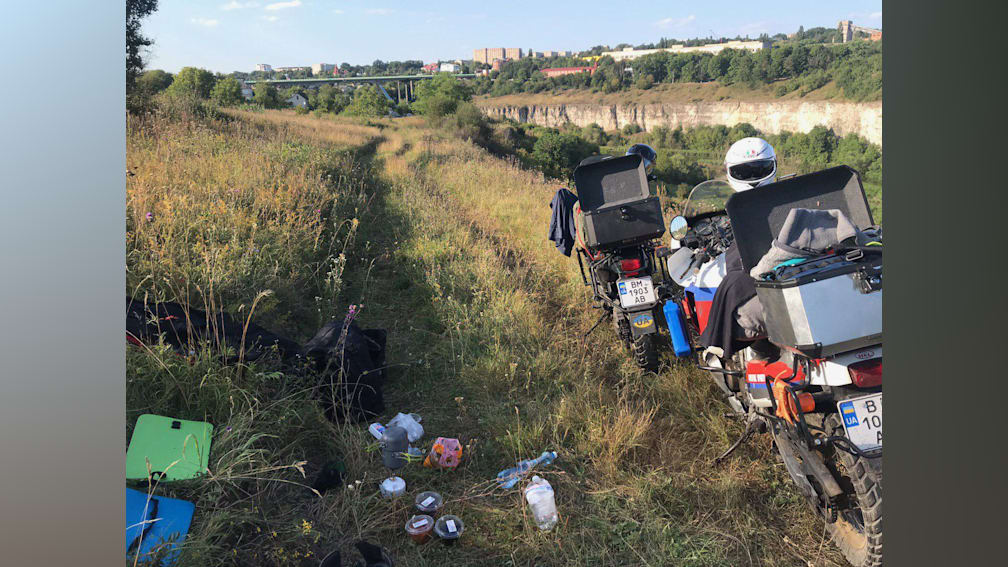A couple of motorcycles are parked on a grassy field with a backpack on the back of one of them.